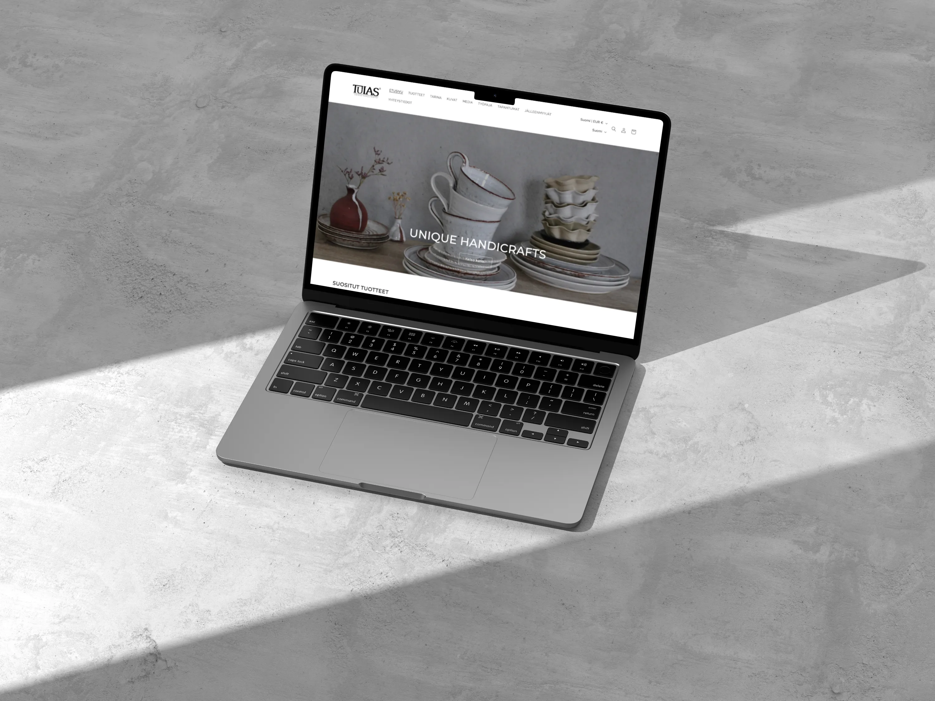 A laptop displays a website featuring handcrafted ceramics, set against a textured gray surface with natural light and shadows.